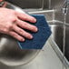 A hand holding a blue Scotch-Brite Low Scratch Scour Pad over a sink.