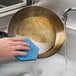 A hand holding a blue Scotch-Brite sponge washing a pan.