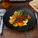 A Tuxton TuxTrendz matte black china plate with a meal of vegetables and meat on a table.