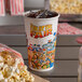 A 16 oz. paper cup with a "Fun at the Fair" design filled with soda and ice with a straw and popcorn.