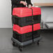 A woman using a dolly to move stacked red and black Metro Mightylite BigBoy food pan carriers.