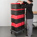 A woman using a dolly to transport a stack of red and black Metro Mightylite pan carriers.