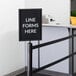 A Lancaster Table & Seating black stanchion sign frame with a clear cover and a sign that says "line forms here"