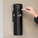 A person in a black jacket uses a Lavex black wall mounted cigarette receptacle to dispose of a lit cigarette.