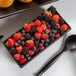 A black rectangular bowl filled with berries with a black spoon next to it.