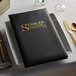 A black Oakmont menu cover with gold corners on a plate with a fork and knife.