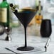 A close-up of a black Stolzle Glisten martini glass with a gold rim filled with a blue liquid and olives.