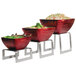 A Vollrath brushed stainless steel square buffet riser with three red bowls of food on it.