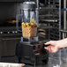A person pouring food into a Vitamix blender on a counter in a professional kitchen.