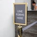 A Lancaster Table & Seating gold stanchion sign frame with a sign with white text on a pole.