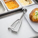 A stainless steel Vollrath ice cream scoop with a baked potato on a plate.