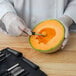 A person in a white coat using a spoon to cut a melon on a wooden surface.