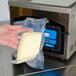 A hand holding a plastic ARY VacMaster vacuum packaging bag with a piece of cheese inside.