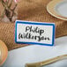A blue ceramic table tent sign with a white border and black text on a table with a name tag that says Philip Wilkinson.
