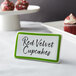 A green ceramic table tent sign with a decal border on a table that says "red velvet cupcakes"