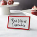 A maroon ceramic table tent sign with a decal border on a table near red velvet cupcakes.