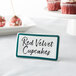 A teal ceramic table tent sign with a decal border on a table next to a red velvet cupcake.
