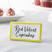 A yellow ceramic table tent sign with a black border and the words "Red Velvet Cupcakes" on it.