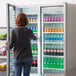 A woman standing in front of a Beverage-Air Marketeer series glass door refrigerator full of drinks.