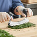 A person using a Mercer Culinary mezzaluna to chop herbs on a table.
