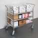 A Regency stainless steel three shelf utility cart with white boxes with black text on them.