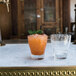 A clear Arcoroc double old fashioned glass filled with orange liquid on a marble counter with another drink and a clear glass.
