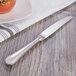 A silver Oneida Baguette stainless steel dessert knife on a table next to a plate of food.