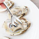 A hand holding a Oneida Baguette stainless steel oyster fork over an oyster shell.