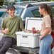A man and woman standing on the back of a truck next to a white CaterGator outdoor cooler filled with cans of soda.