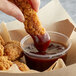 A hand dipping a fried chicken strip into a cup of Sweet Baby Ray's Honey BBQ Sauce.