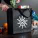 A person putting food into a small black LK Packaging reusable shopping bag with a white sun design on it.