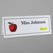 A clear plastic tent card holder holding a white card with black text and a red apple with a green leaf.