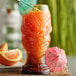 A Head Hunter tiki glass filled with an orange drink and decorated with umbrellas and an orange slice.