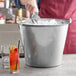 A person stirring ice in a Vollrath stainless steel dairy bucket filled with drinks.