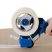 A hand using a blue and white Shurtape General Purpose masking tape dispenser to seal a cardboard box.