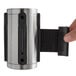 A hand holding a CSL silver stainless steel stanchion head with a black retractable belt.