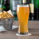 A Libbey pilsner glass of beer on a coaster on a table with a bucket of fries.