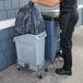 A man holding a black garbage bag next to a Rubbermaid Slim Jim trash can.