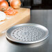 An American Metalcraft Super Perforated Heavy Weight Aluminum Coupe Pizza Pan on a counter next to onions and a cutting board.