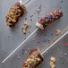 A chocolate covered banana with sprinkles and nuts on a Paper Chocolate Stick.