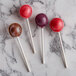 A hand holding a white Paper Lollipop Stick next to three round lollipops with red, purple, and brown tops on a marble surface.