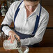 A woman in a white shirt and Henry Segal navy suspenders pouring a drink into a clear glass with ice.