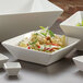 A close-up of a white square bowl with a salad in it.