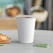 A white Choice paper hot cup filled with coffee sits on a table next to a croissant.