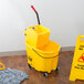 A Rubbermaid yellow mop bucket with a side press wringer and mop on a wood floor with a yellow caution sign.