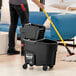 A person pushing a Rubbermaid black mop bucket with wheels.