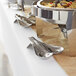 A Tablecraft brushed stainless steel double spoon rest holding a spoon and fork on a buffet table.