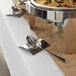 A Tablecraft brushed stainless steel square spoon rest holding a spoon on a buffet table.
