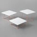 A white square table with copper wire legs holding a rose gold Acopa square plate riser set.