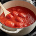 A #10 can of Conte whole peeled plum tomatoes in puree with a wooden spoon in a pot of red sauce.